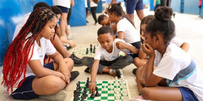 Group of children sitting on the floor, playing a chess game on a large board in a school hallway or gym area, focused and chatting.