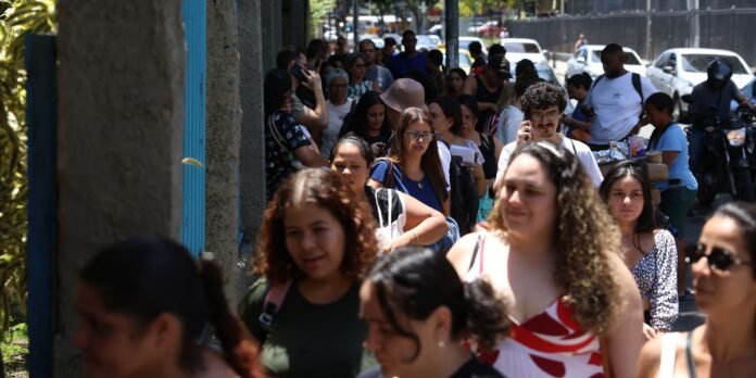 Crowded city sidewalk with diverse pedestrians walking in the sun, many looking at phones, forming a long queue along the street.