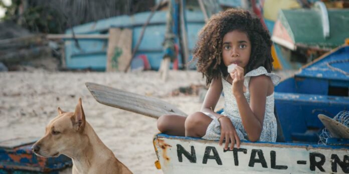 Girl with curly hair sits in a small, weathered boat on a sandy beach, eating ice cream while a dog stands nearby in the foreground, colorful boats in the background.