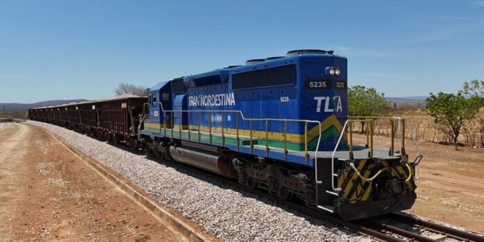 Blue diesel locomotive pulling a long line of brown freight cars on a curved gravel railway in a dry, rural landscape under a clear blue sky.