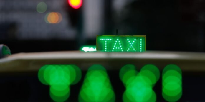 Green illuminated taxi sign on the roof of a car at night, with bokeh green street lights in the foreground and blurred background.