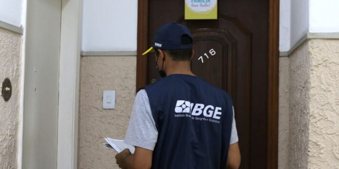Male field worker in a navy IBGE vest and cap, standing with papers at a doorway.
