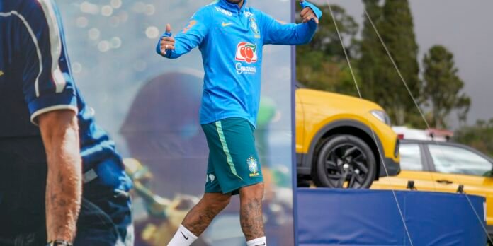 Brazilian footballer in a blue training kit gives thumbs up; tattoos on the legs, yellow car in the background.