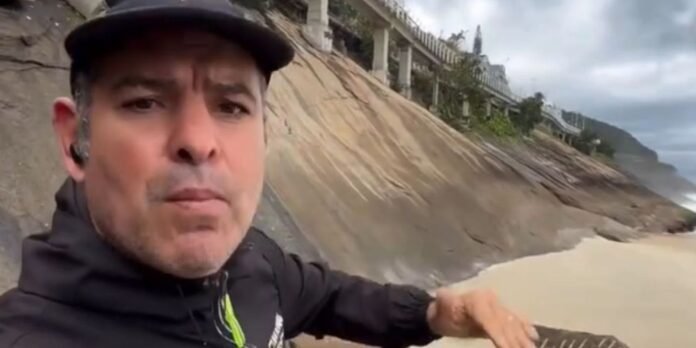 Man in a black jacket and cap selfie-filming on a sandy beach, with a steep rocky wall and houses along the cliff behind him.