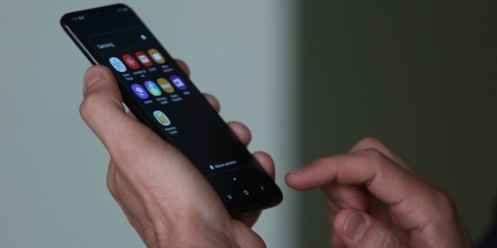 Person holding a smartphone at an angle, displaying app icons on the home screen on the device's dark screen.