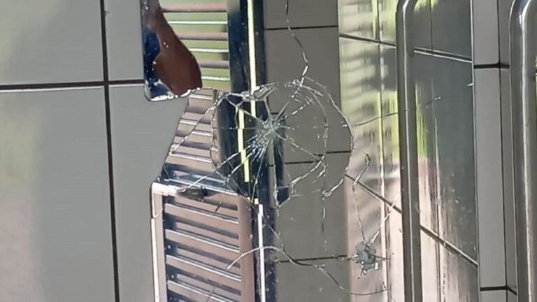Circular spiderweb crack on a glass pane with radiating fractures near a metal frame inside a building.