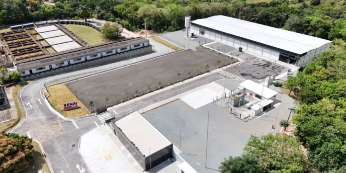 Aerial view of an industrial campus with white-roofed buildings, a large paved lot, and surrounding trees; a curved road leads to the entrance with a Senai sign nearby.