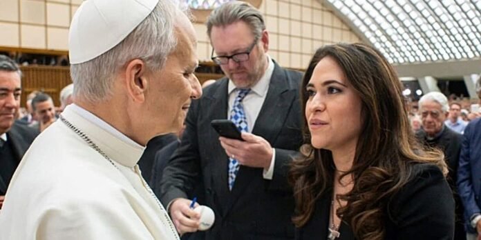 Pope greets a woman in a black blazer at a crowded indoor event, a man in a suit records with a smartphone in the background.