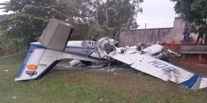 Small single-engine airplane wreckage on grassy area with debris scattered and two police officers in the background near a concrete wall, after a crash.