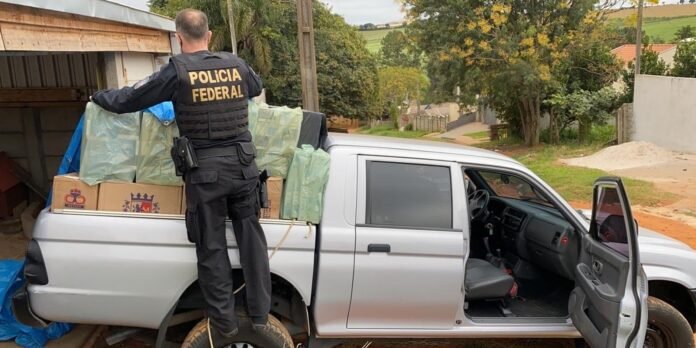 Polícia Federal officer loading plastic-wrapped boxes into the back of a silver pickup truck in a residential area, wearing a vest that reads 'POLICIA FEDERAL'.