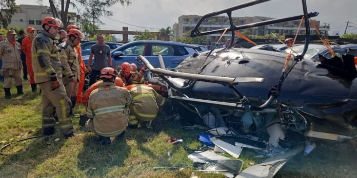Firefighters in orange and tan gear work at the wrecked black helicopter on a grassy area as bystanders watch nearby.