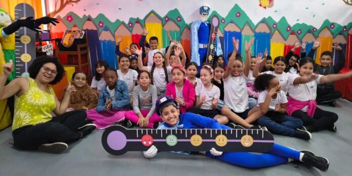 Group of smiling students sitting in a circle on the floor, posing with a large mood-meter prop and a teacher on the left in a colorful classroom.