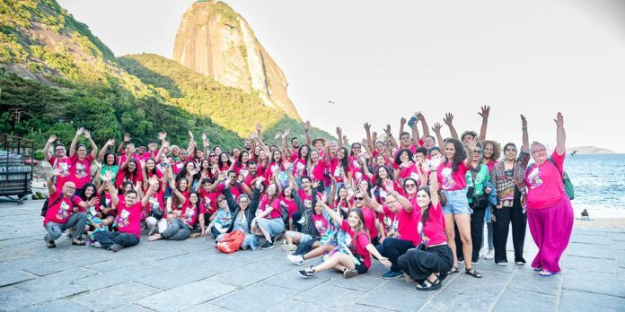 Large group of people wearing matching pink shirts pose with raised hands on a stone seaside promenade, with a rocky hill and the ocean in the background.
