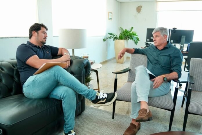 Two men discuss in a bright office; one sits on a green sofa with a clipboard, the other on a gray chair with papers in hand.