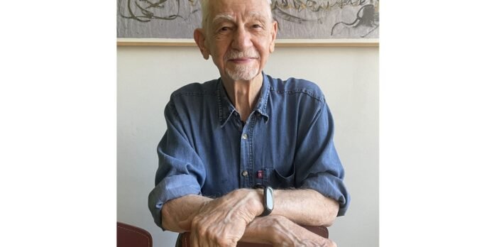 Elderly man with white hair and goatee sits at a table with arms crossed, wearing a denim shirt and a smartwatch, in a light room.