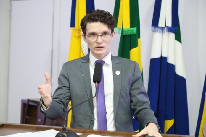 Man in a gray suit and purple tie speaks at a podium with a microphone, flags in the background.