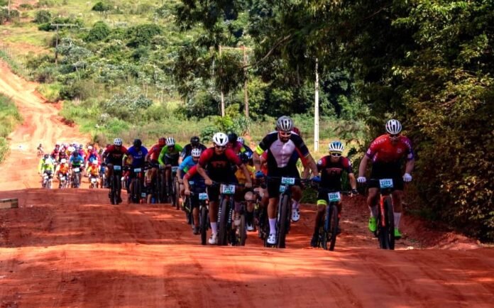 A large group of cyclists wearing colorful jerseys race along a dusty red dirt road surrounded by green hills and trees.