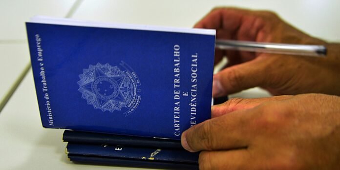 Blue Brazilian employment and social security booklet (Carteira de Trabalho e Previdência Social) being held in hands, with a stack nearby.