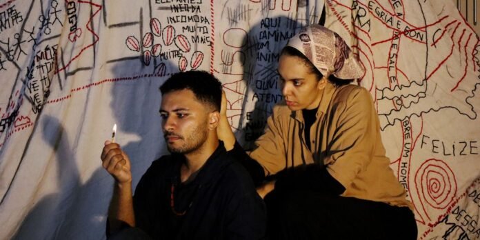 Two people sit against a large mural with red lines and text; the man looks at a lit match he holds while the woman beside him rests her hand on his shoulder, watching closely.