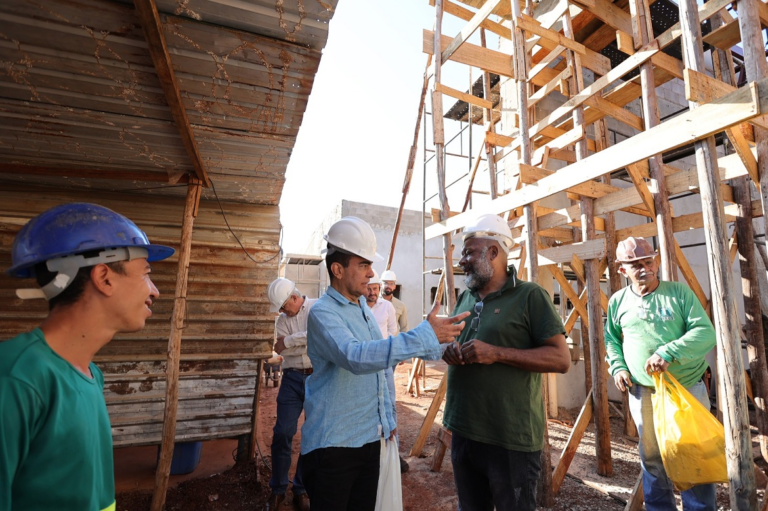 Group of construction workers wearing hard hats stand among wooden scaffolding; two men in conversation in the center of the site.