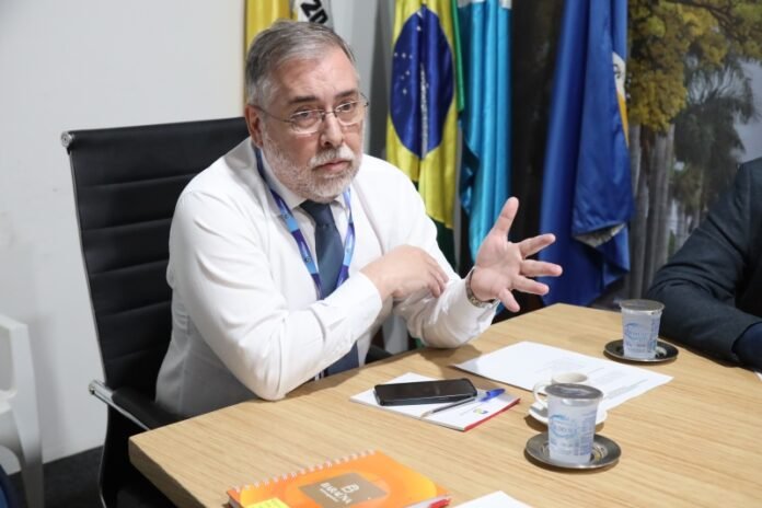 Man with gray hair and beard in a white shirt and tie explains something at a meeting, gesturing with his hands; flags in the background behind him.