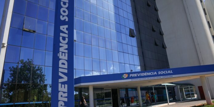Modern glass-fronted office building with a tall blue banner reading PREVIDÊNCIA SOCIAL beside the entrance; pedestrians walk into the doorway.