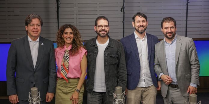 Group of five professionals posing in a studio behind two microphones, smiling at the camera.