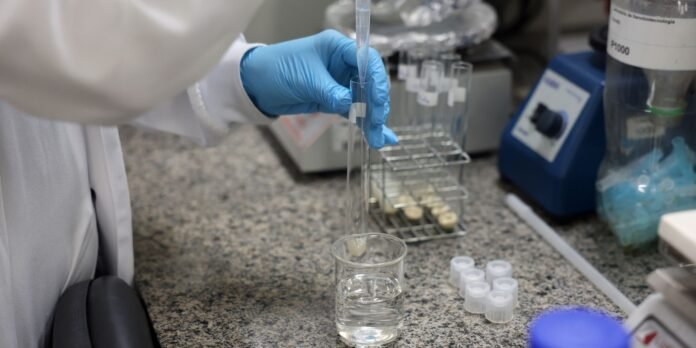 Scientist wearing blue gloves uses a pipette to transfer liquid into a tall test tube on a lab bench.