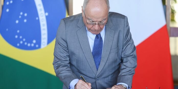 Man in a gray suit signing a document at a formal event, with Brazil and France flags in the background.
