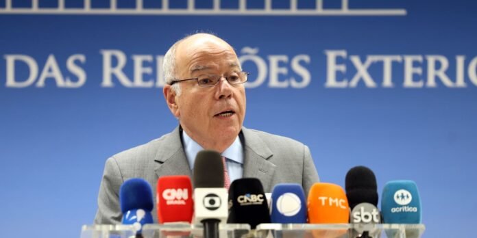 Older man in a gray suit speaking at a press conference, with multiple colorful microphones in front.