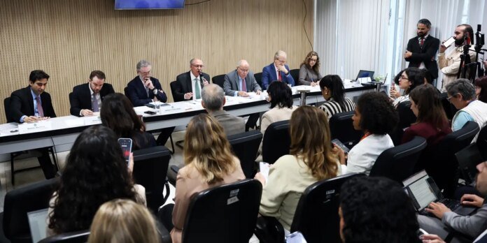 Panel of professionals at a long table speaking to an audience in a conference room; photographers and journalists in the back, attendees using laptops and phones.