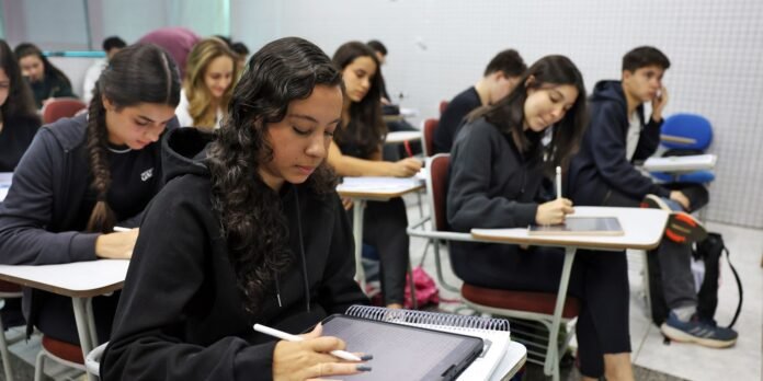 Group of students in a classroom taking notes, using tablets and notebooks on their desks.