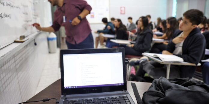 Instructor writes on a whiteboard as students sit and take notes in a classroom; a laptop is open on the foreground desk.