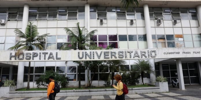Facade of a university hospital with a large 'HOSPITAL UNIVERSITARIO' sign, glass windows, and palm trees; two pedestrians with backpacks walk by.