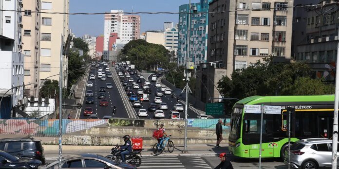 Busy urban street scene with a multi-lane highway, a green city bus on the right, and cyclists and pedestrians at a crosswalk surrounded by tall buildings and graffiti walls.