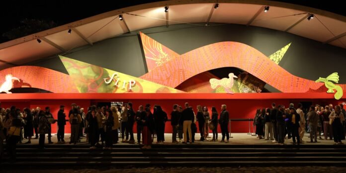 Nighttime scene of a crowd lined up along a red wall under a curved, colorful mural at a gallery entrance or event venue.