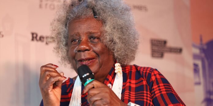 Elderly Black woman with gray curly hair speaking into a handheld microphone, wearing a red plaid blouse and white beaded necklace at an event.
