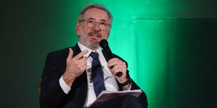 Older man in a dark suit and blue tie speaks into a handheld microphone on a green-lit stage.