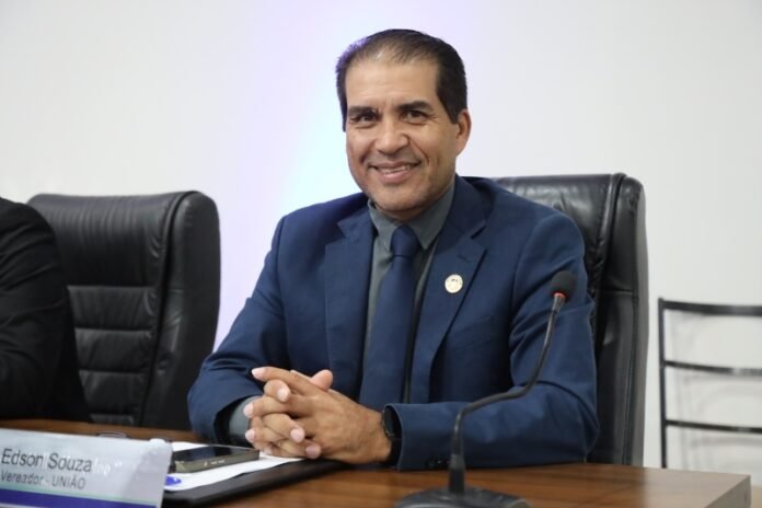 Man in a dark blue suit and tie, seated at a conference table with a microphone, smiling at the camera.