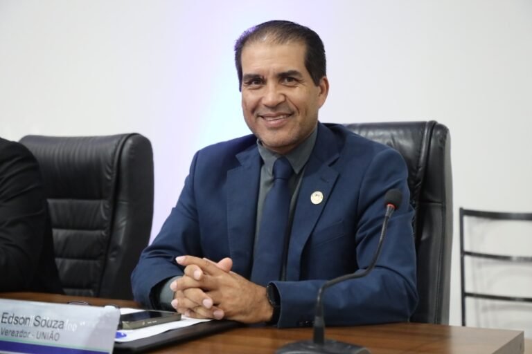 Man in a dark blue suit and tie, seated at a conference table with a microphone, smiling at the camera.