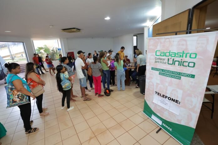 People line up at a registration desk in a bright interior, a large 'Cadastro Único' banner visible nearby.
