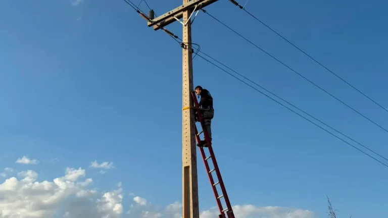 Worker on a red ladder repairing overhead power lines on a wooden utility pole against a blue sky.