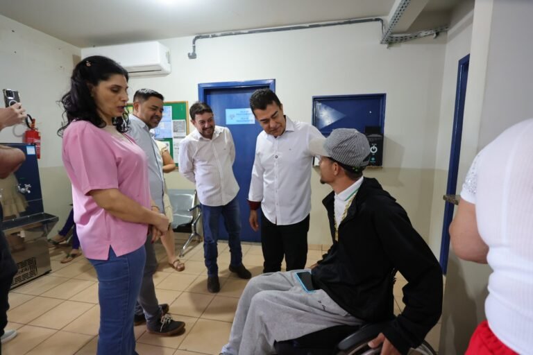 Group of adults standing around a man in a wheelchair, indoors, chatting in a clinic or hospital hallway.