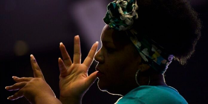 Side-profile of a woman with a colorful headwrap, raising hands in a performative gesture against a dark background