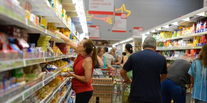 Woman in a red sleeveless top reaches for items on a grocery store shelf while other shoppers push carts in the background.