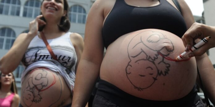 Pregnant woman in a black top with a cartoon sheep drawn on her belly in marker, a smiling friend in a white tank top nearby.