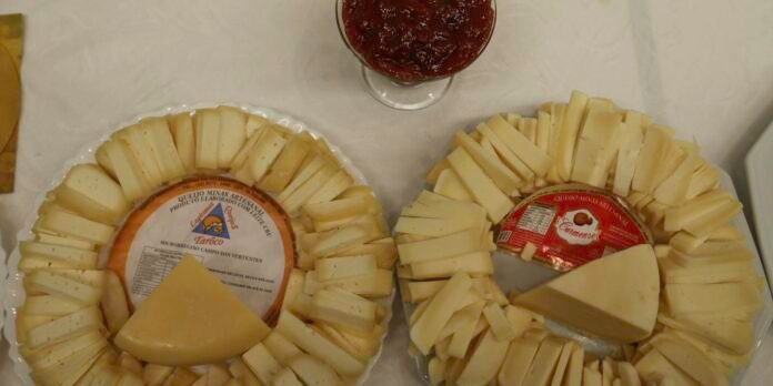 Two circular cheese wheels ringed with sliced cheese on white platters. A wedge has been cut from each wheel. A jar of red jam sits above.