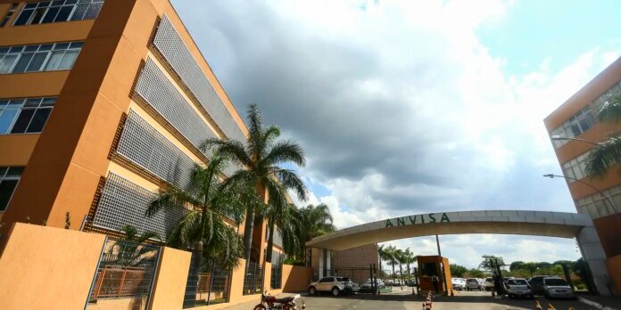 Exterior view of an orange building complex with a white archway labeled ANVISA, palm trees, and parked cars at the entrance side.