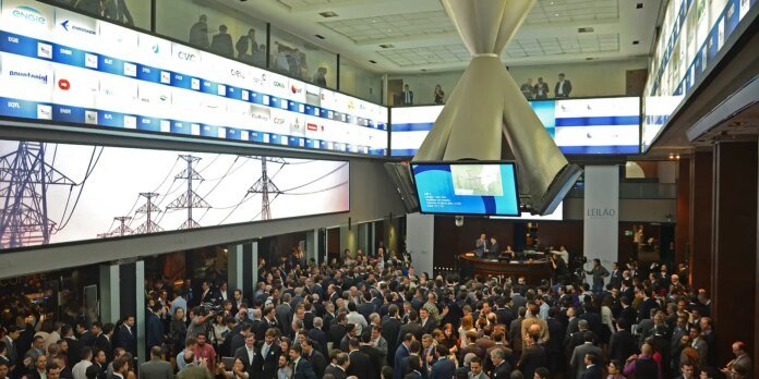 Large crowd of suited professionals at a busy conference or exhibition in a modern atrium with screens and a central monitor display.