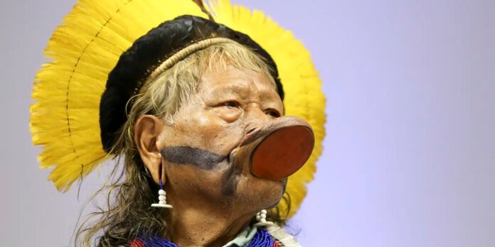 Person wearing a large prosthetic nose, bright yellow feather headdress, blue beads, and a dark black stripe across the cheek.